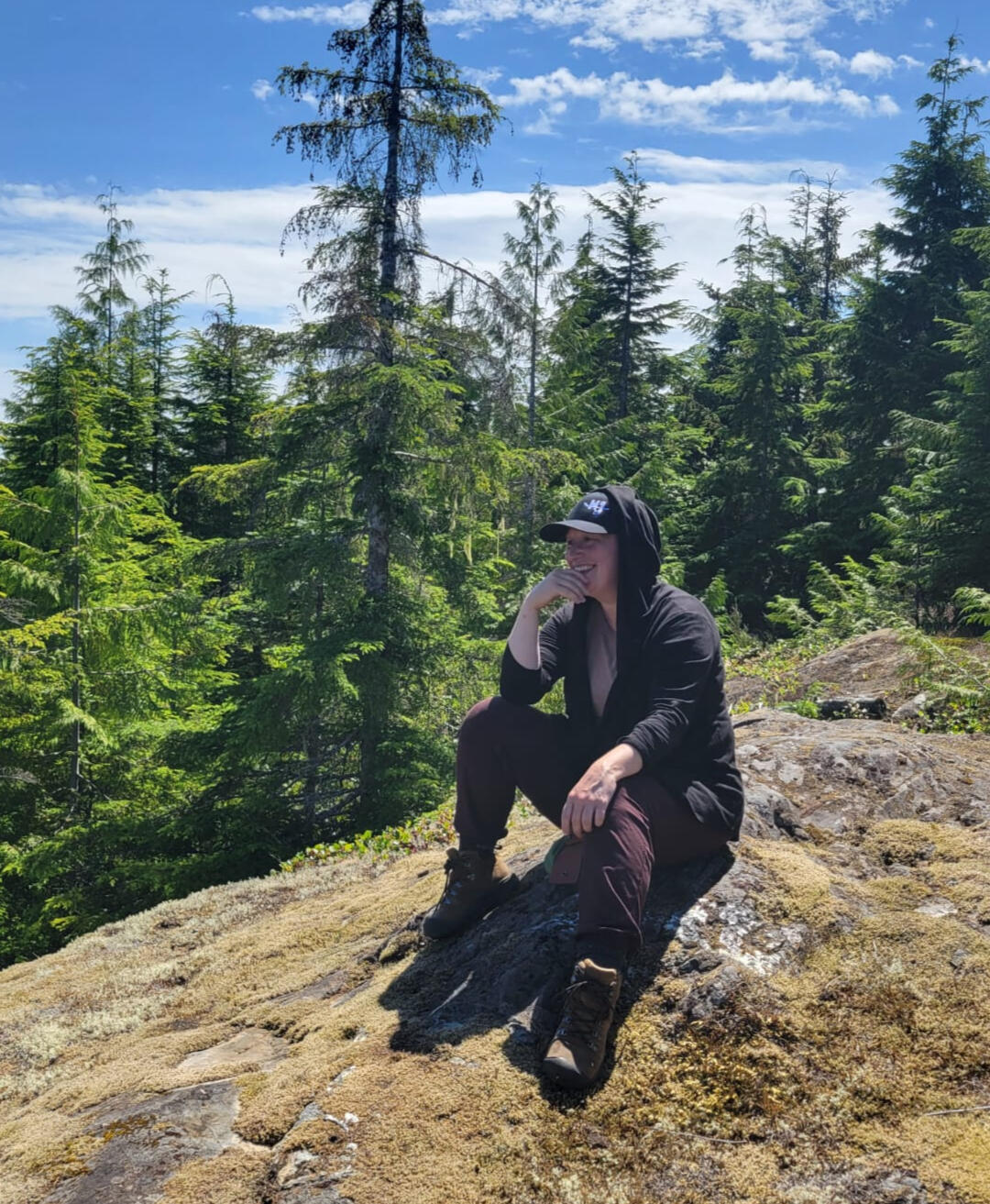 Jaymi sitting on a rocky outcrop surrounded by evergreen forest.