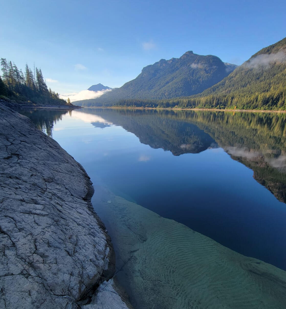 Mountain and forest reflected in still blue water beside a rocky shoreline.
